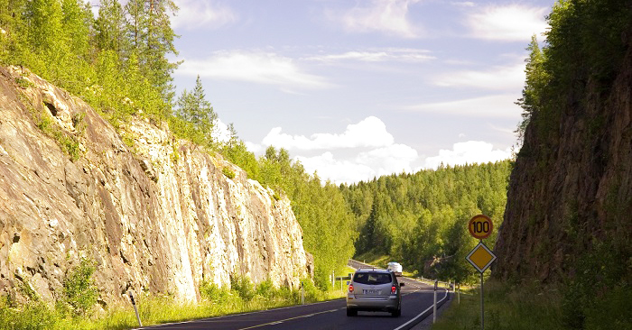 Auto ajaa tietä, missä on kallioleikkaus ja sadan kilometrin nopeusrajoitusmerkki. 